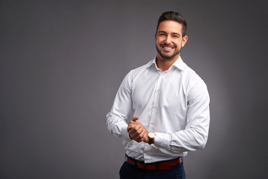 A handsome confident young man standing and smiling in a white shirt.